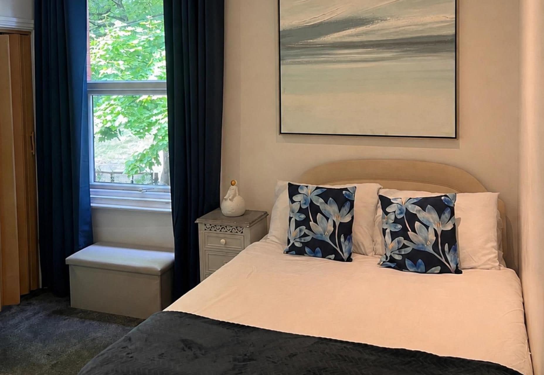 A bedroom with a bed, a nightstand, a window with curtains, and a painting on the wall.