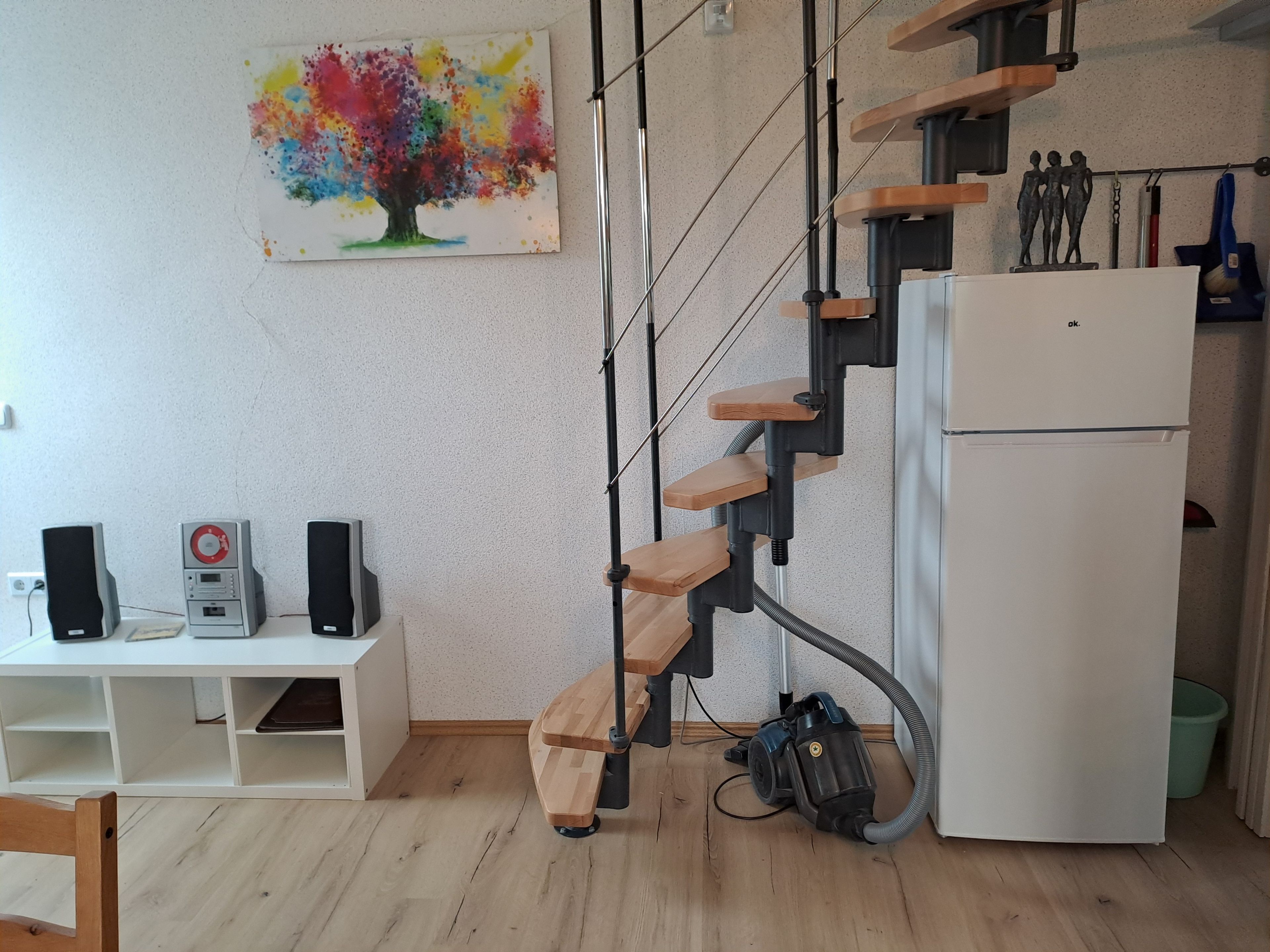 Een keuken met een witte koelkast, een houten trap en een schilderij van een boom.