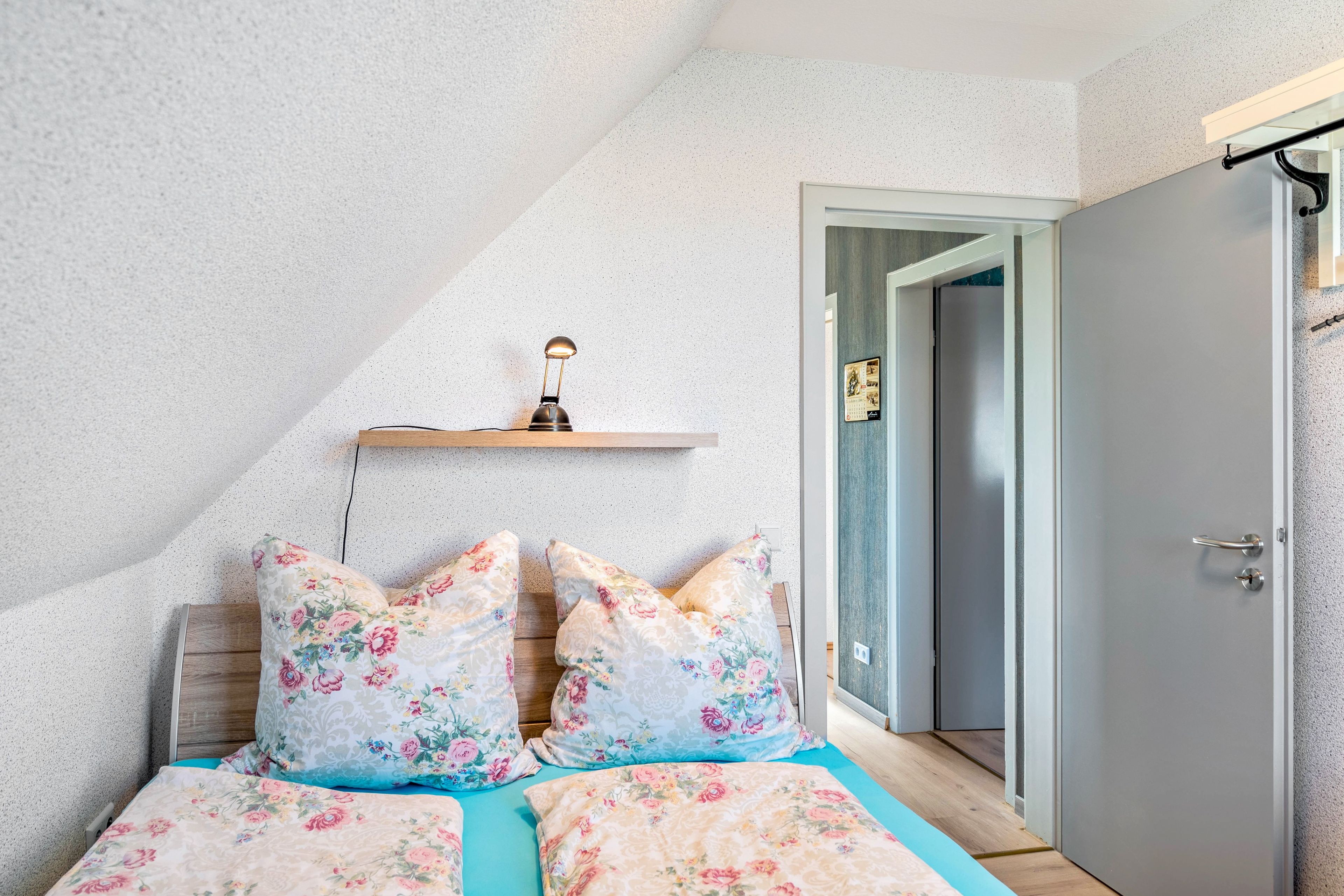 A bedroom with a bed, pillows, a nightstand, a wall-mounted lamp, and a door.