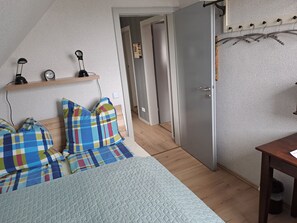 Quarto com cama, mesa de cabeceira, abajur, relógio e estante com objetos decorativos.