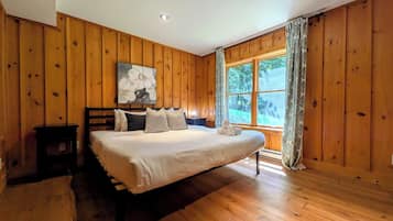 A bedroom with a large bed, wooden walls, a window with curtains, and a painting on the wall.