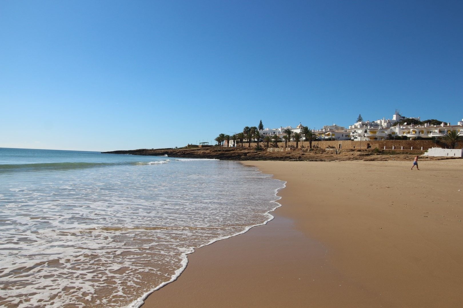 A beach with a clear sky, a sandy shore, and buildings in the background.