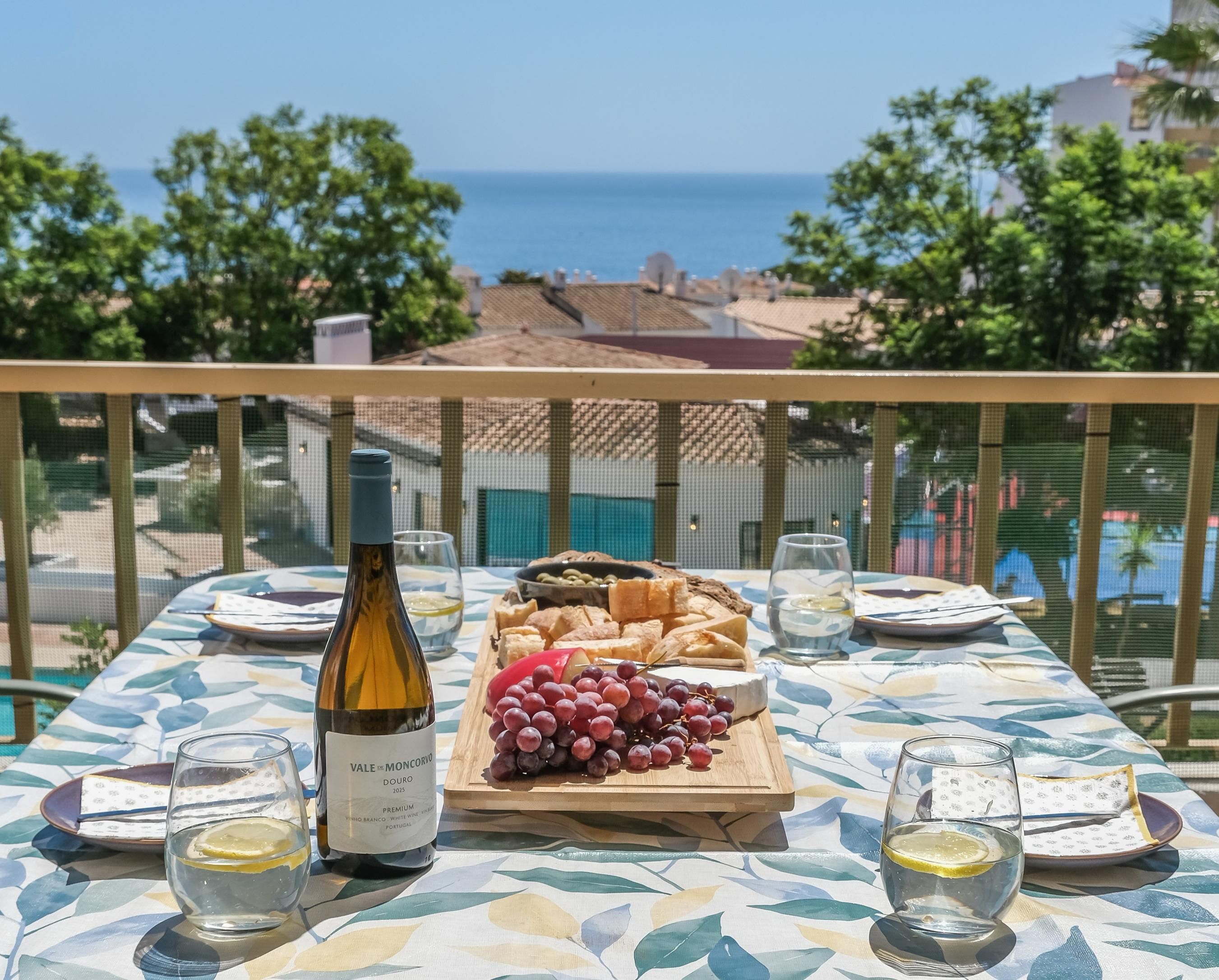 A table set with a bottle of wine, a wooden board with cheese and grapes, and glasses of water, overlooking a coastal view.