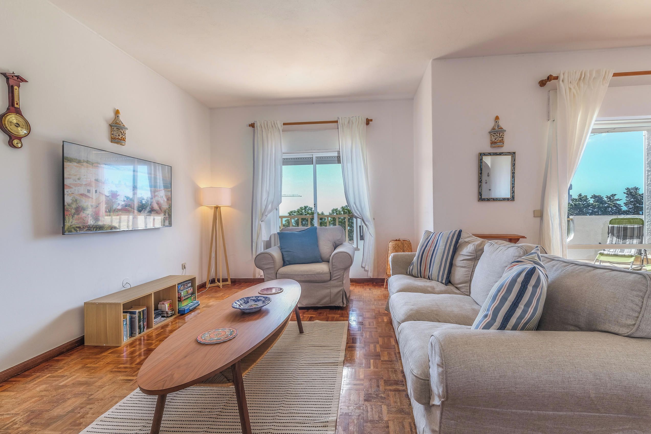 A living room with a sofa, a table, and a chair. There is a window with curtains, a lamp, and a bookshelf.