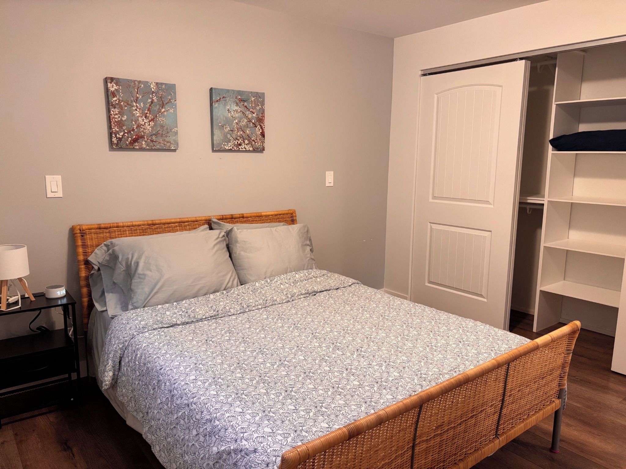 A bedroom with a bed, a nightstand, a lamp, a wall-mounted shelf, and a wardrobe.