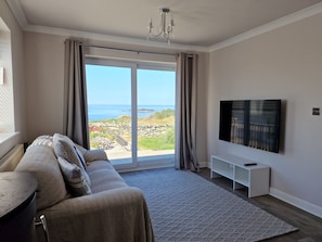 A modern living room with a sofa, a flat-screen TV, a large window with a view of the sea, and a chandelier.