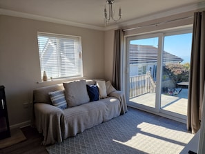 A room with a sofa, a chandelier, a sliding glass door leading to a balcony, and a window with blinds. - Seaside Bungalow with Wide Reaching Views (Amlwch)