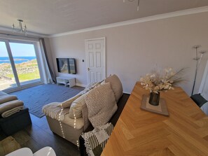 A living room with a sofa, a wooden table, and a view of the outdoors. - Seaside Bungalow with Wide Reaching Views (Amlwch)