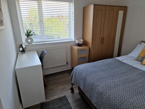 A bedroom with a bed, a wardrobe, a desk, a chair, a radiator, and a window with blinds. - Seaside Bungalow with Wide Reaching Views (Amlwch)