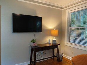 A room with a flat-screen TV mounted on the wall, a small table with a lamp, a potted plant, and a window with blinds. - Cozy 1-bedroom Cottage-In-The-Trees!. (Tallahassee)