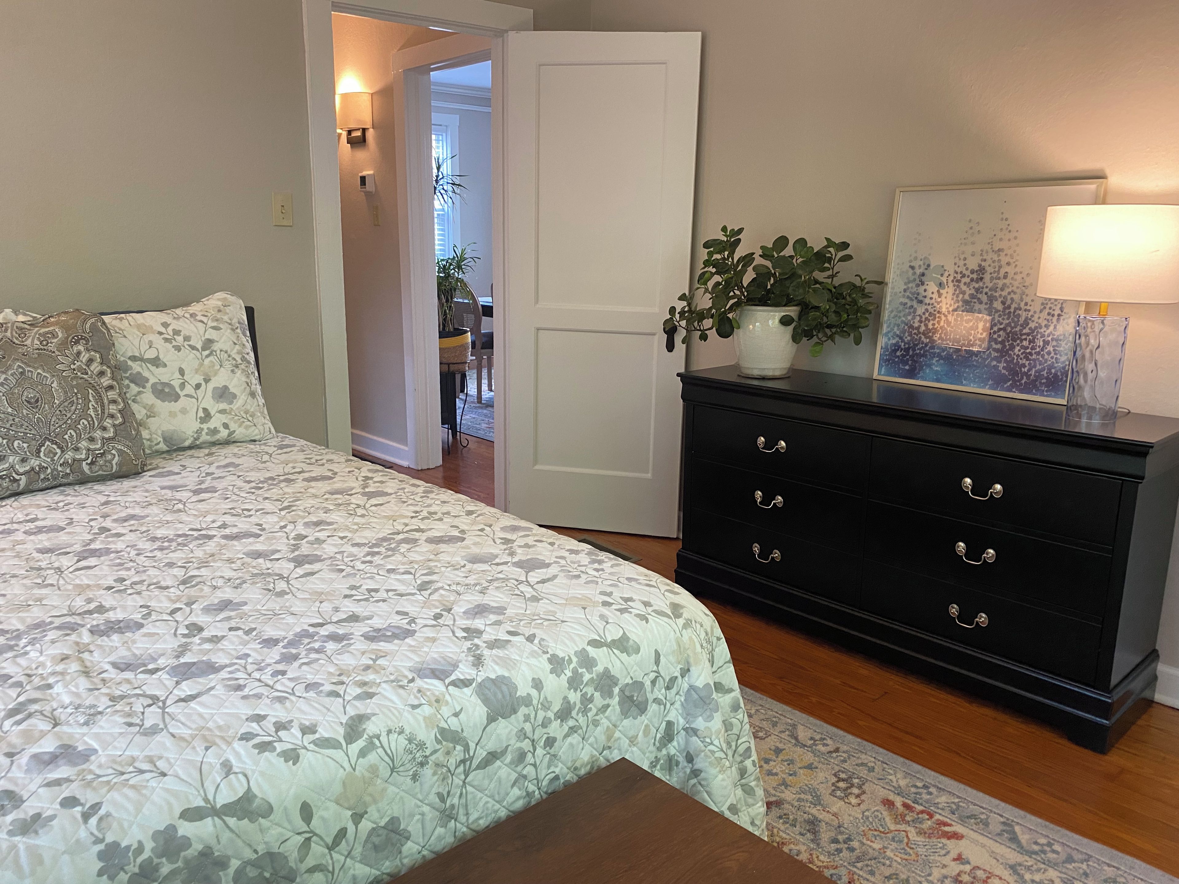 A bedroom with a bed, a dresser, a plant, and a lamp.