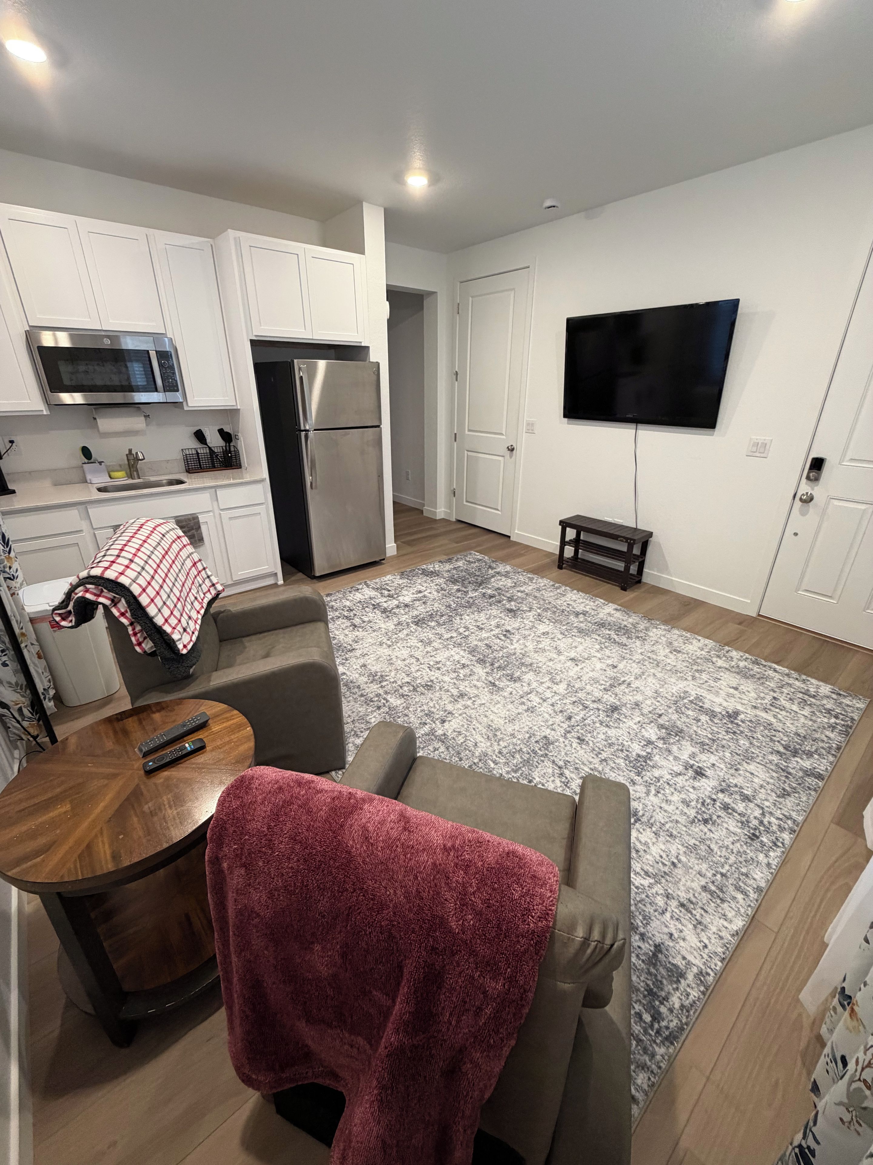 A modern kitchen with white cabinets, a microwave, and a refrigerator. There is a gray rug on the floor, a small wooden table with two remotes, and a gray sofa with a red blanket. A flat-screen TV is mounted on the wall.