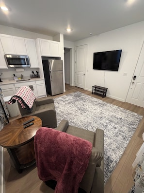 Een moderne keuken met witte kasten, een magnetron en een koelkast. Op de vloer ligt een grijs vloerkleed, bij een klein houten tafeltje liggen twee afstandsbedieningen, en er is een grijze bank met een rode plaid. Een flatscreen televisie hangt aan de muur.