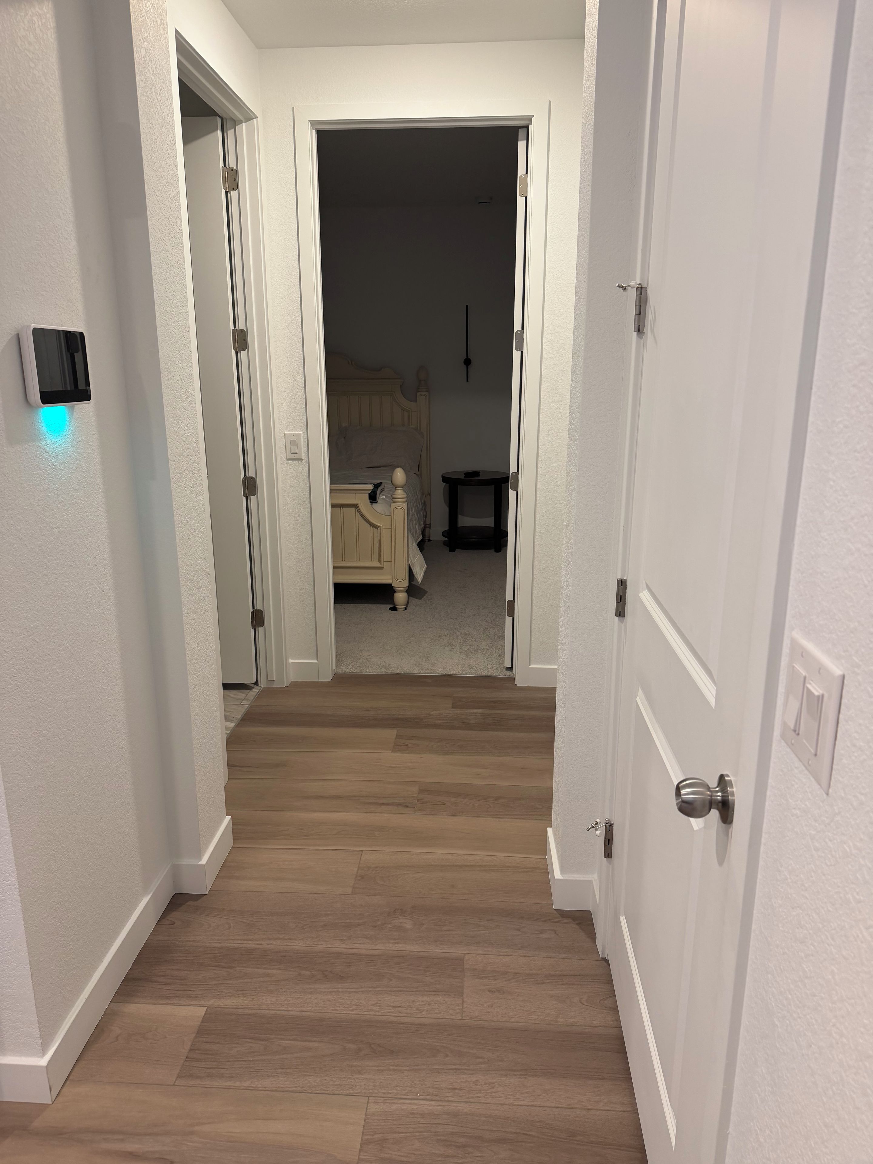 A hallway leading to a bedroom with a bed, bedside table, and a wall-mounted light fixture.