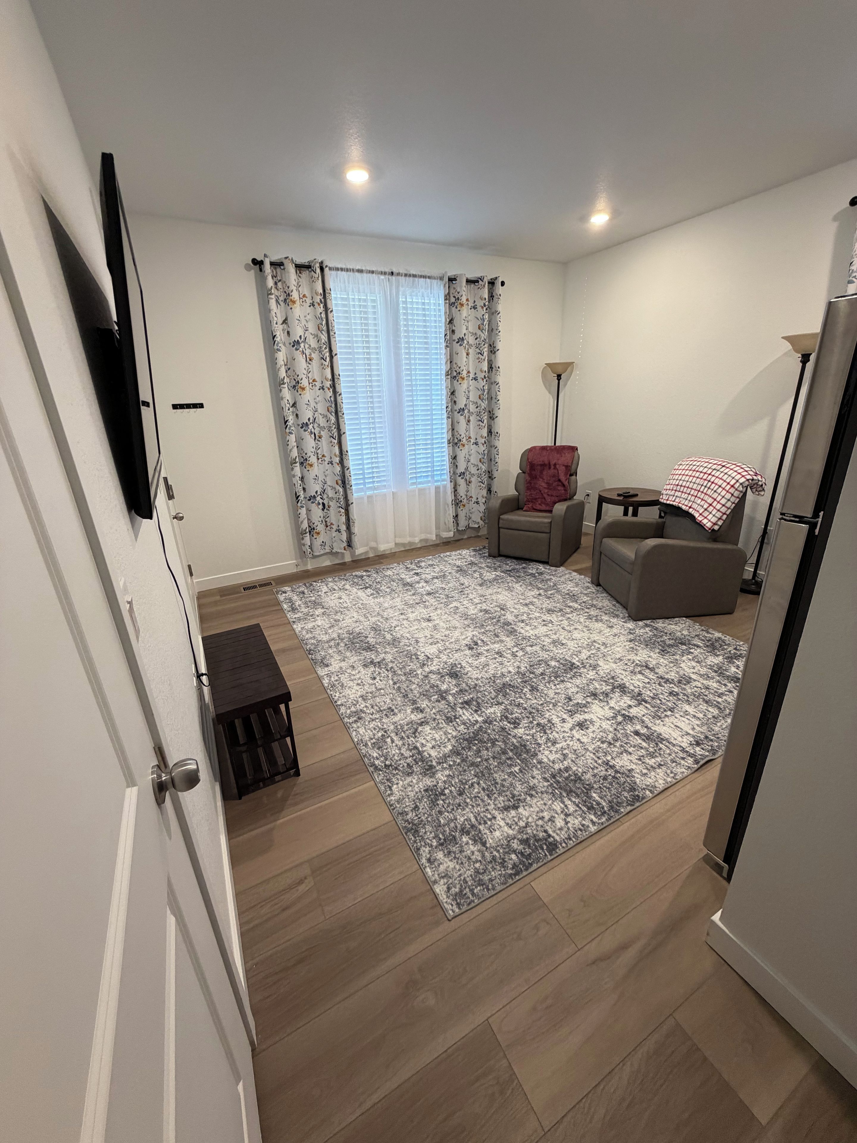 A living room with a grey patterned rug, a sofa, a chair, a small table, and a television mounted on the wall.