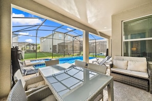 A covered outdoor area with a glass table and chairs, a sofa, and a view of a swimming pool and houses. - Sleeps 15·Private Pool·Themed Rooms·Game Room (Kissimmee)