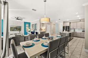 A modern kitchen with a central island, a dining area with a long table, and a living area with a sofa and TV. - Sleeps 15·Private Pool·Themed Rooms·Game Room (Kissimmee)