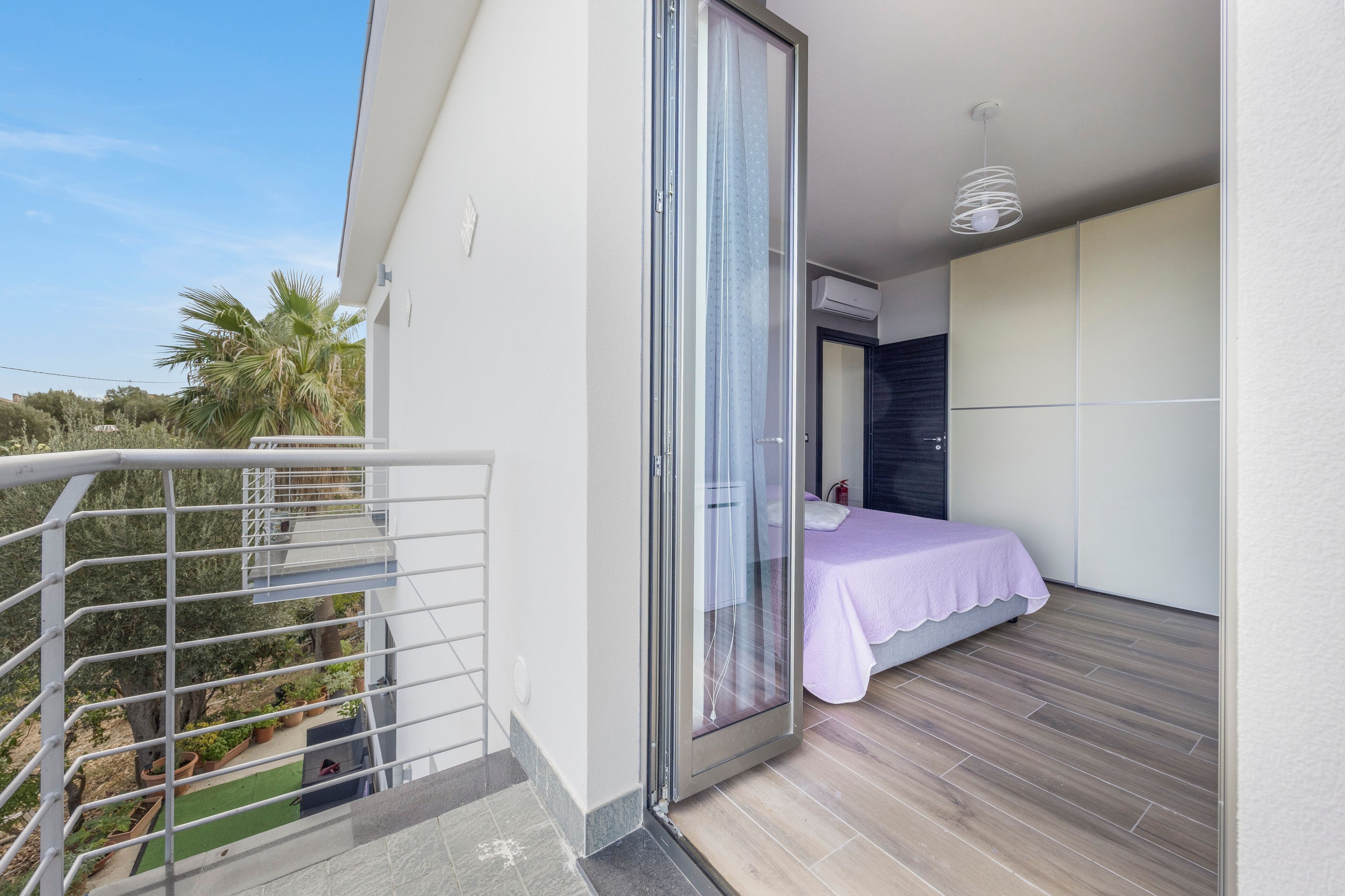 Ein Balkon mit Metallgeländer, ein Bett mit weißer Bettdecke und Blick auf Palmen und Grünpflanzen.
