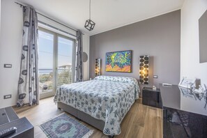 A bedroom with a bed, a nightstand, a desk, a chair, a window with curtains, a painting on the wall, and a view of the outdoors.