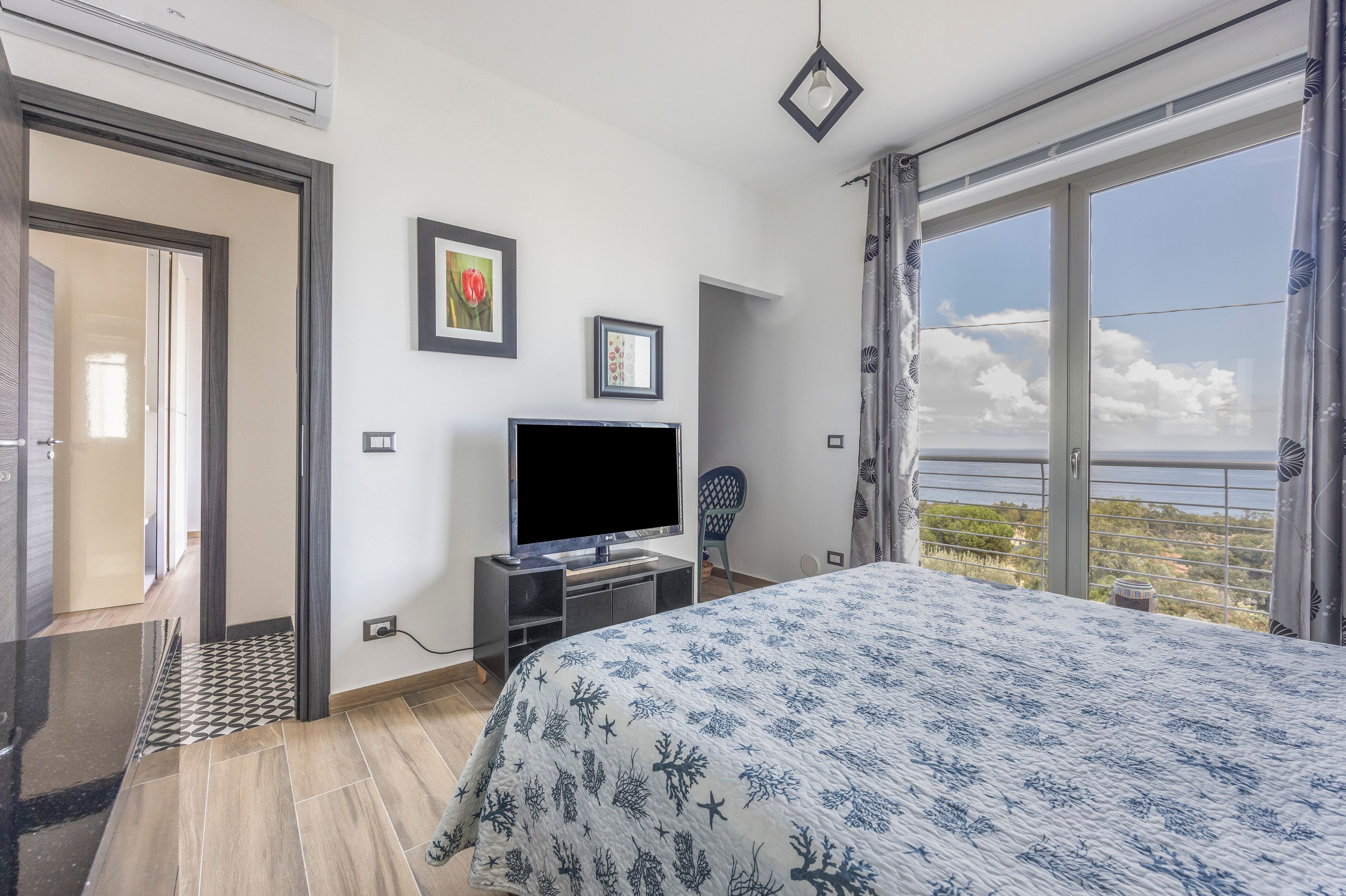 A hotel room with a bed, a TV, a door, and a view of the sea.