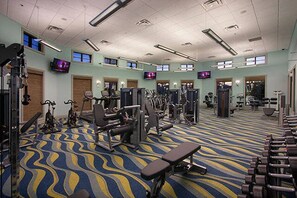 Ein Fitnessraum mit unterschiedlichen Trainingsgeräten, einem gemusterten Teppich und mehreren Fernsehbildschirmen an den Wänden.