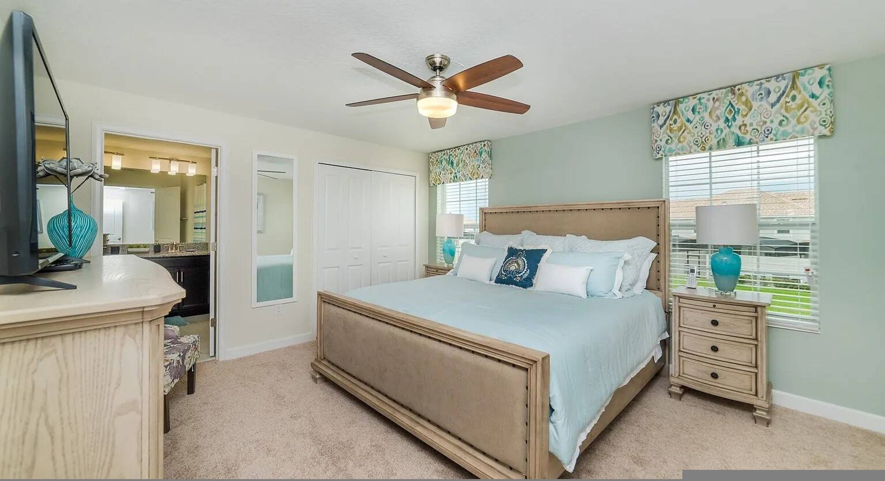 A bedroom with a bed, bedside tables, a ceiling fan, a dresser, a mirror, and a window with blinds.