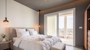 Un dormitorio con una cama grande, mesitas de noche, un banco, un ventanal con vistas y una puerta que da a un espacio exterior.