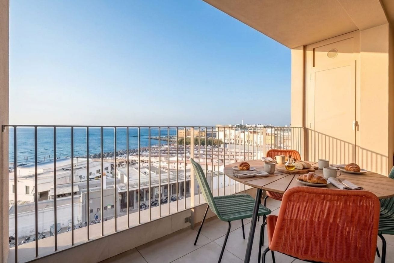 Un balcon avec vue sur la mer et les bâtiments, une table avec des éléments pour le petit-déjeuner et deux chaises.