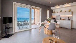 Una sala de estar moderna con un televisor de pantalla plana, un comedor con mesa y sillas, una cocina con armarios y encimera, y un amplio ventanal corredero que da a un balcón con vistas al mar.