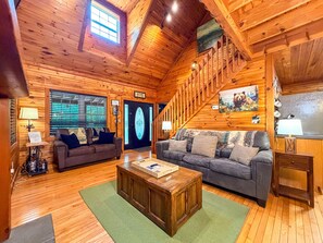 A log cabin interior with a wooden staircase, two sofas, a coffee table, and a welcome sign. - Cozy 2BR/2BA Helen Cabin for 4 | Hot Tub & Firepit (Sautee Nacoochee)