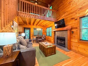 Interior de una cabaña de troncos con sala de estar, comedor, chimenea y un balcón.