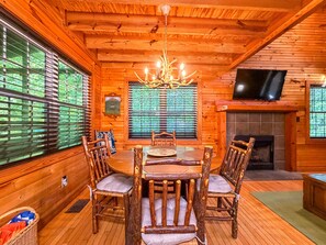 Interior de una cabaña de madera con una mesa de comedor, sillas, una araña, chimenea y televisor.