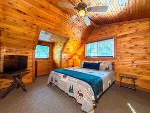 A cozy log cabin bedroom with a bed, a TV, a window, and a ceiling fan. - Cozy 2BR/2BA Helen Cabin for 4 | Hot Tub & Firepit (Sautee Nacoochee)