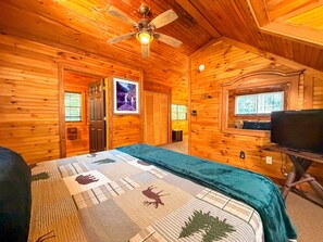 Una habitación de cabaña de madera con una cama, un ventilador de techo, un televisor y una ventana.