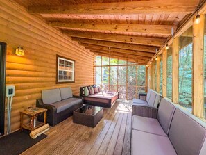 A log cabin interior with a wooden deck, a sofa, a table, and a window overlooking a forest. - Cozy 2BR/2BA Helen Cabin for 4 | Hot Tub & Firepit (Sautee Nacoochee)