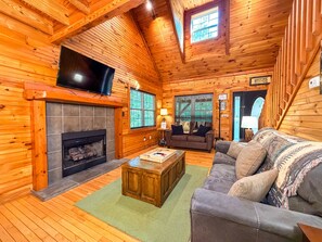 A log cabin interior with a fireplace, a television mounted above it, a sofa, and a coffee table. - Cozy 2BR/2BA Helen Cabin for 4 | Hot Tub & Firepit (Sautee Nacoochee)