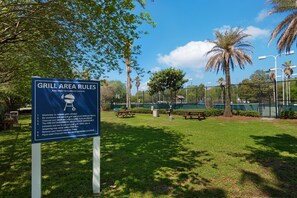 Schild mit Regeln für den Grillbereich in einem Park mit Picknicktischen und Palmen.