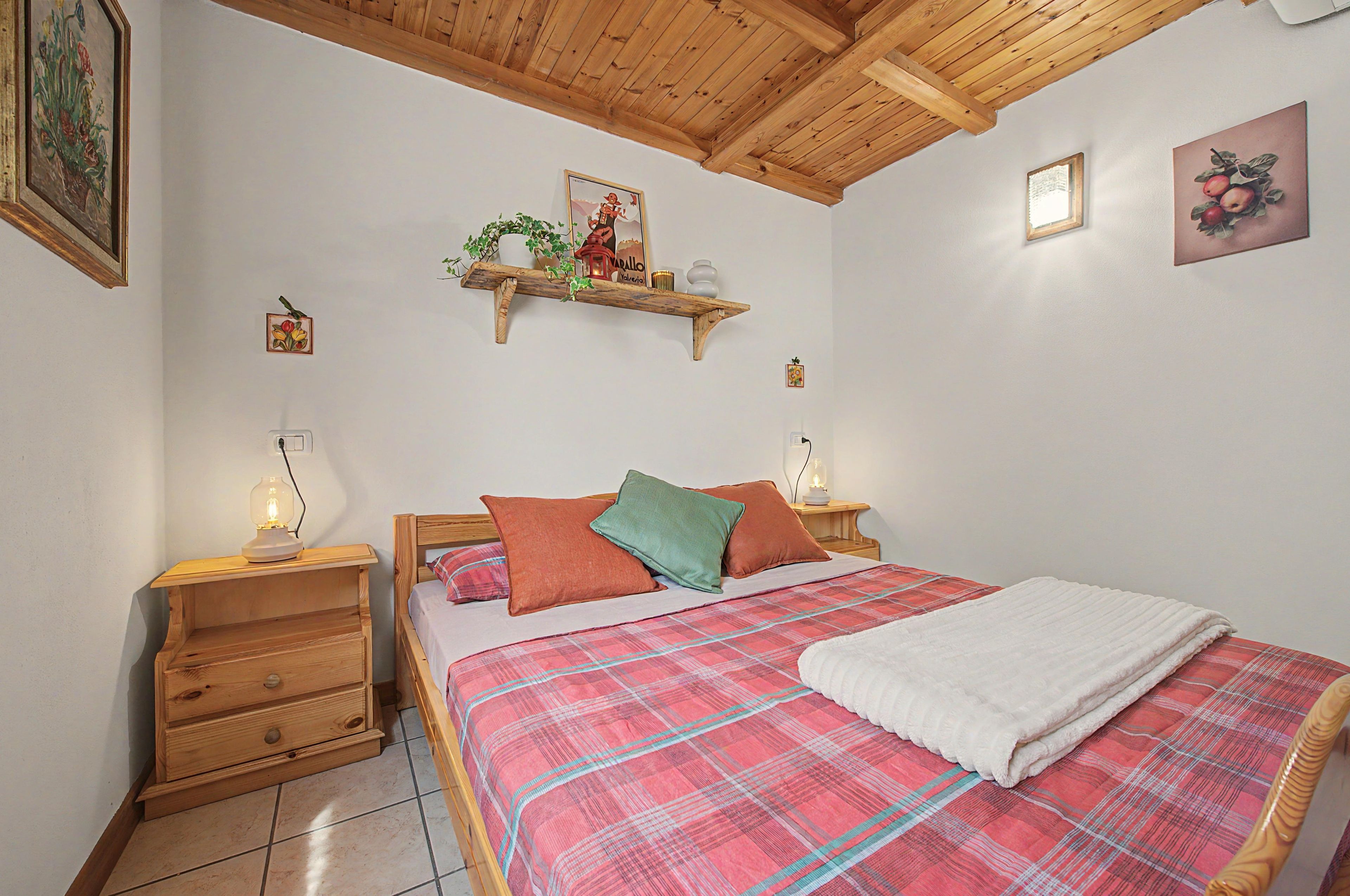 A bedroom with a wooden bed, a nightstand with a lamp, a wooden dresser, and a ceiling with exposed beams.