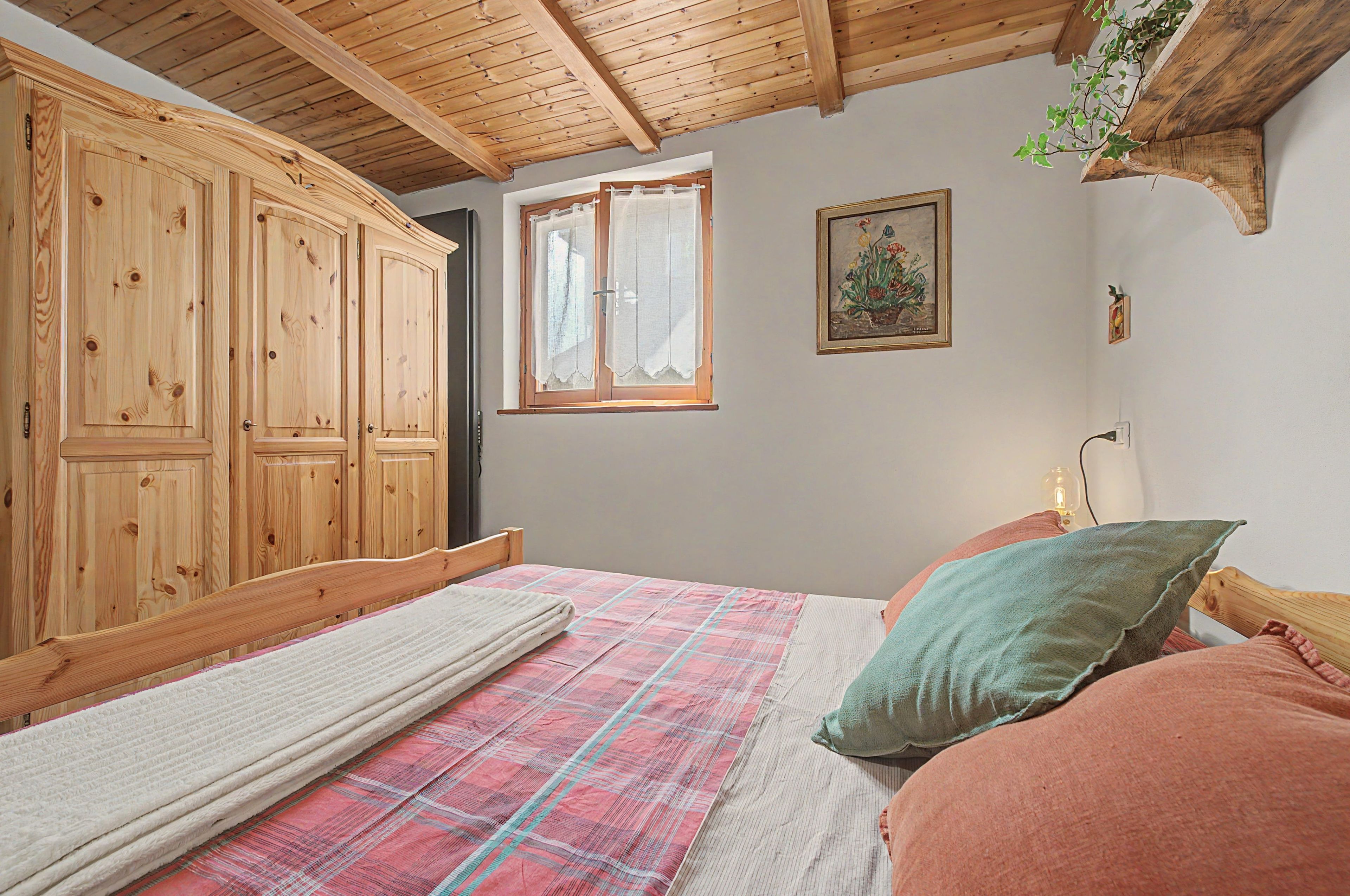 Une chambre avec une armoire en bois, un lit recouvert d’une couverture à carreaux et une fenêtre avec des rideaux.