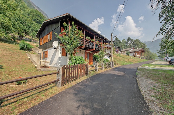 A mountain lodge with a wooden facade, a balcony with potted plants, and a paved driveway leading to the entrance.