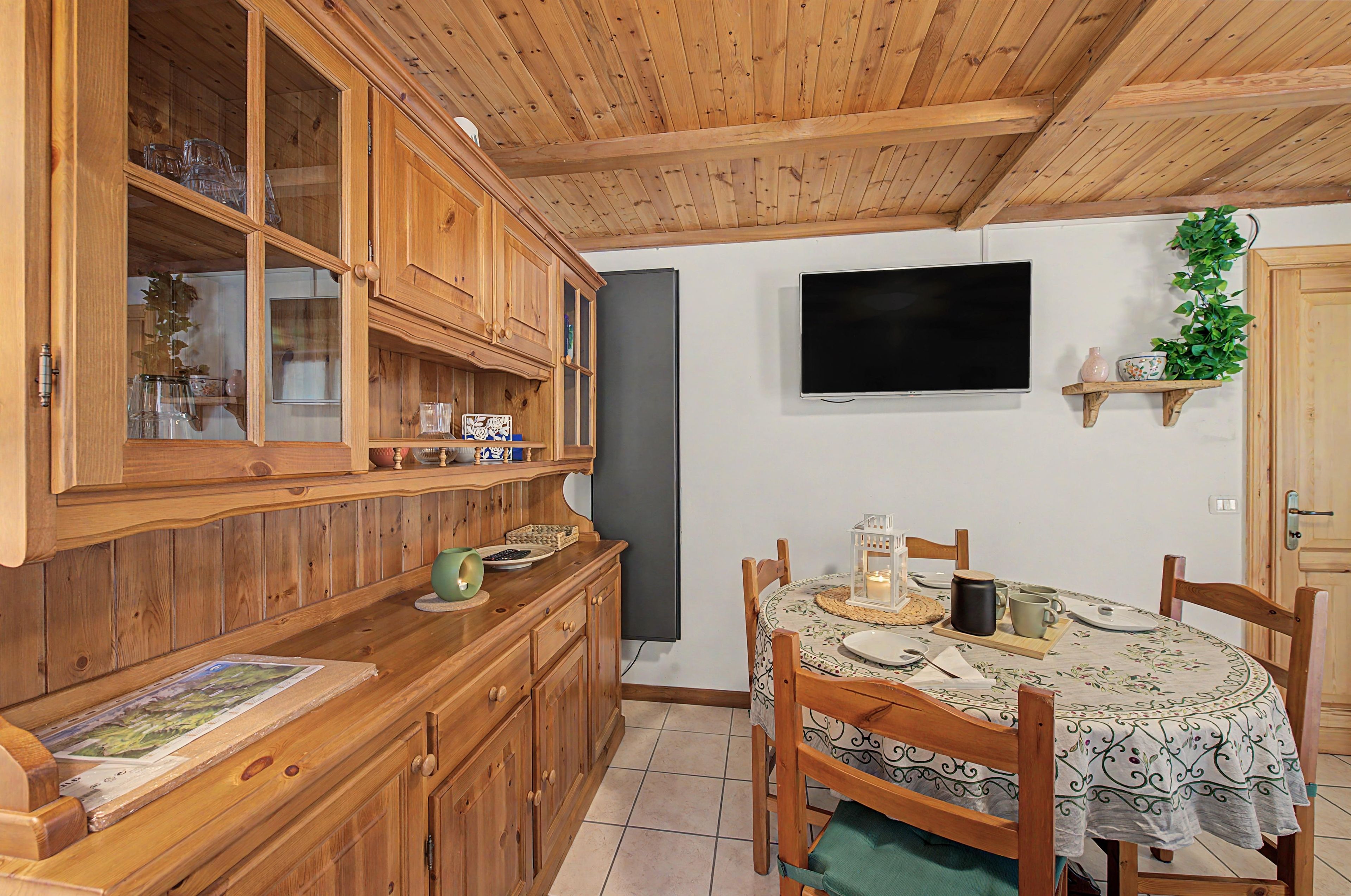 Un comedor de madera con una mesa preparada para dos, un televisor montado en la pared y un mueble con puertas de vidrio.