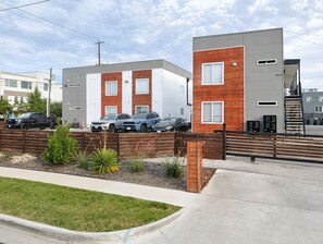 Um edifício residencial moderno com fachada cinza e vermelha, área de estacionamento com vários carros e um jardim bem cuidado com plantas e cerca de madeira.