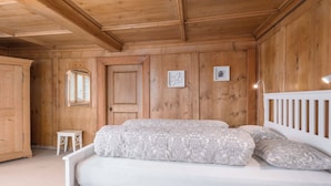 Ein Schlafzimmer mit Holzbalkendecke, einem Bett mit gemusterten Textilien, einem kleinen weißen Nachttisch und einer Tür mit Fenster.