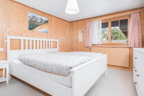 A bedroom with a white bed, wooden walls, a window with curtains, and a painting on the wall.