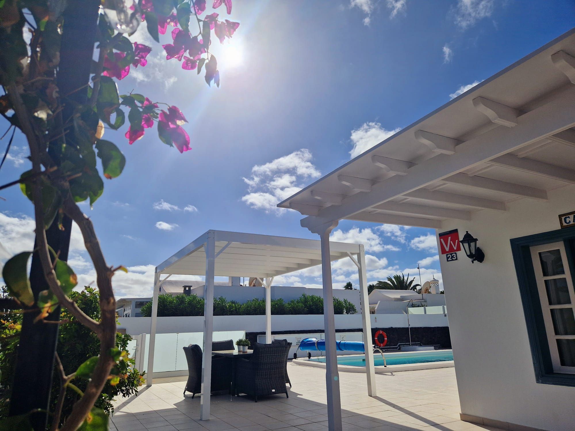 A covered outdoor area with a pool, a table and chairs, and a white building with a sign.