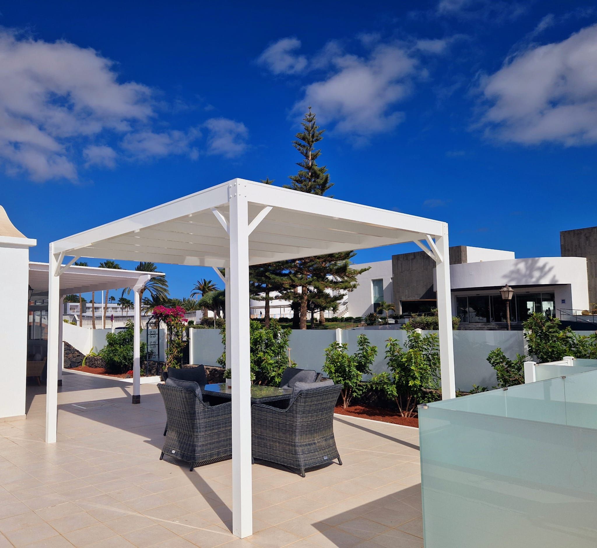 En udendørs patio med hvid pergola, fletmøbler og en klar blå himmel.