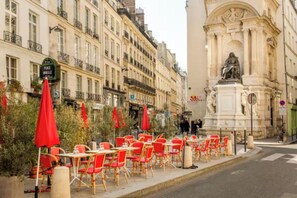 Mobiliário de café ao ar livre com cadeiras vermelhas e guarda-sóis, uma estátua e um edifício com arquitetura clássica.