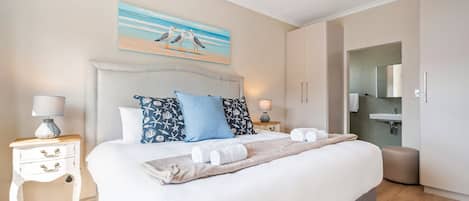 A bedroom with a bed, bedside tables, a painting, a bathroom with a sink, and a mirror.