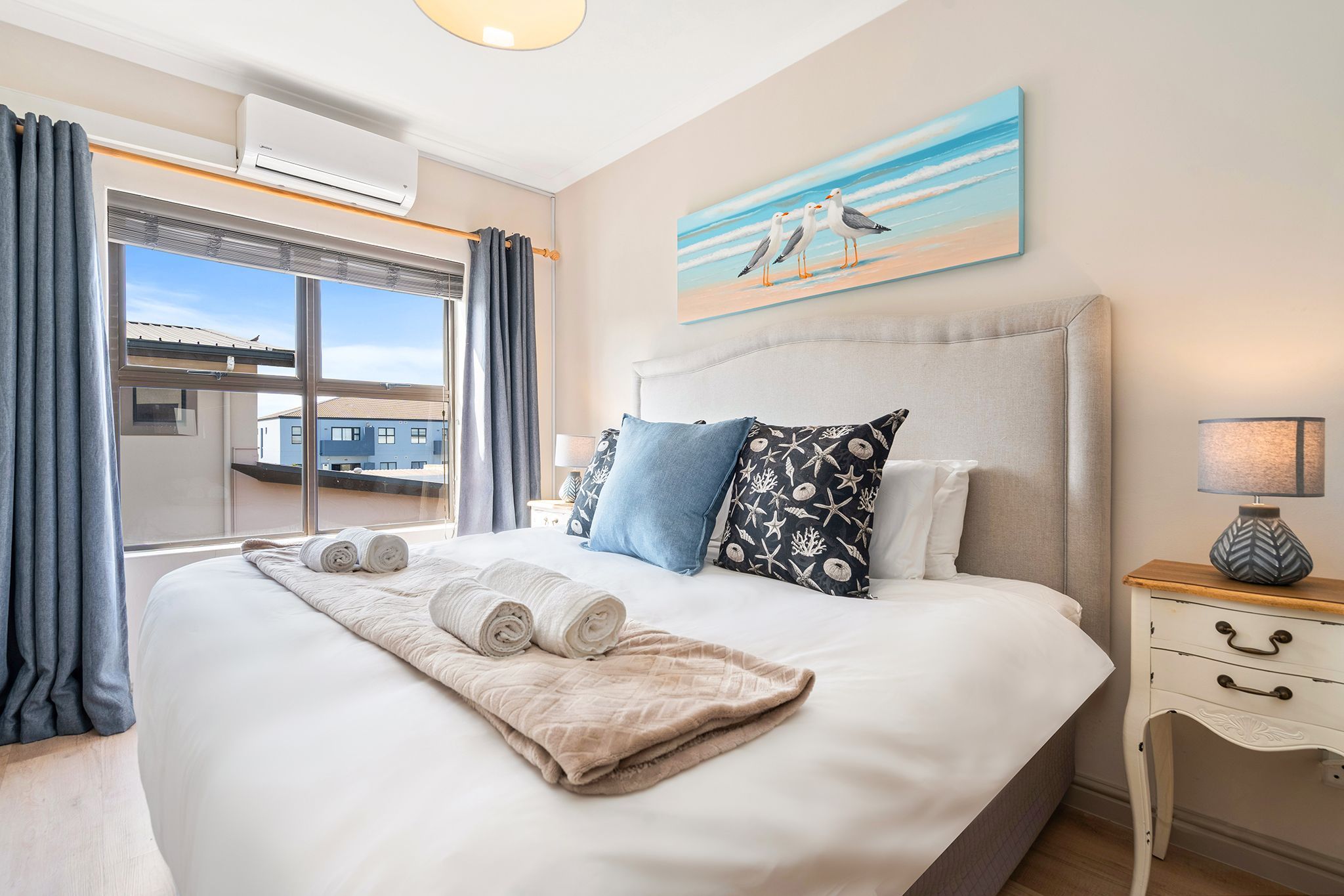 A hotel room with a large bed, a painting of seagulls, a nightstand, and a view of buildings outside.