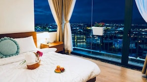 Habitación de hotel con una cama grande, una mesita de noche, una lámpara, una ventana con cortinas y vistas a la ciudad.